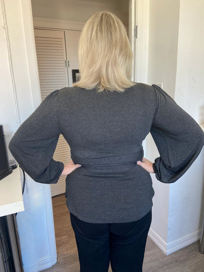 Back view of  woman wearing the Heather Black Bamboo Wrap Top from Dotty, with bracelet length sleeves and adjustable V-neckline. She is wearing it with a black pants and standing in a doorway. 