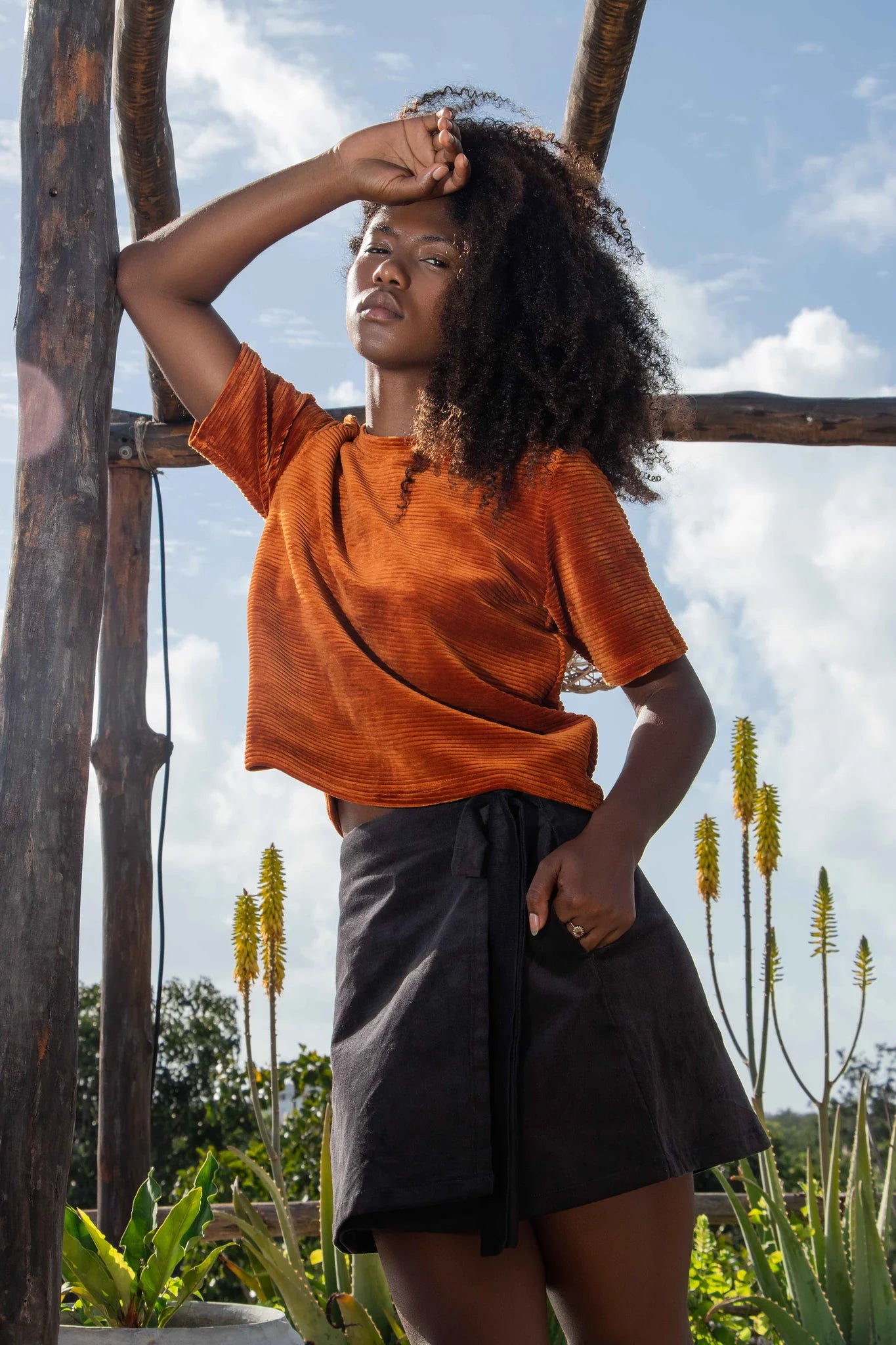 A woman wearing the Tofino Top by MAS in Amber, a slightly cropped tee in velvet rib. She is wearing it with a black skirt and is standing outside. 