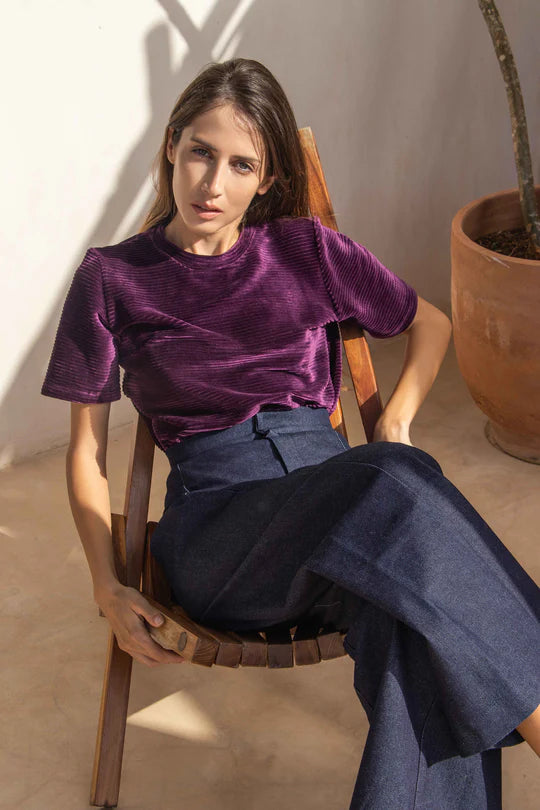 A woman wearing the Tofino Top by MAS in Amethyst, a slightly cropped tee in velvet rib. She is wearing it with a blue trousers and is sitting in a wooden chair. 