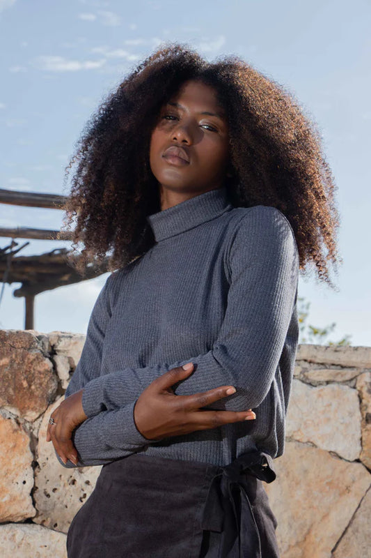 Waist up view of a woman wearing the Tucson Top by MAS in Iron with a dark skirt, standing outside on front of a rocky wall 