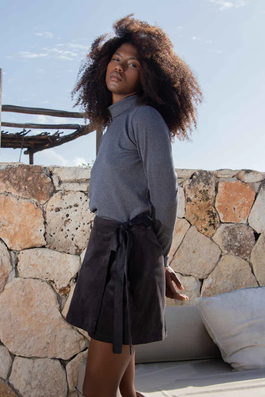 3/4 view of a woman wearing the Tucson Top by MAS in Iron with a dark skirt, standing outside in front of a rocky wall and a low sofa