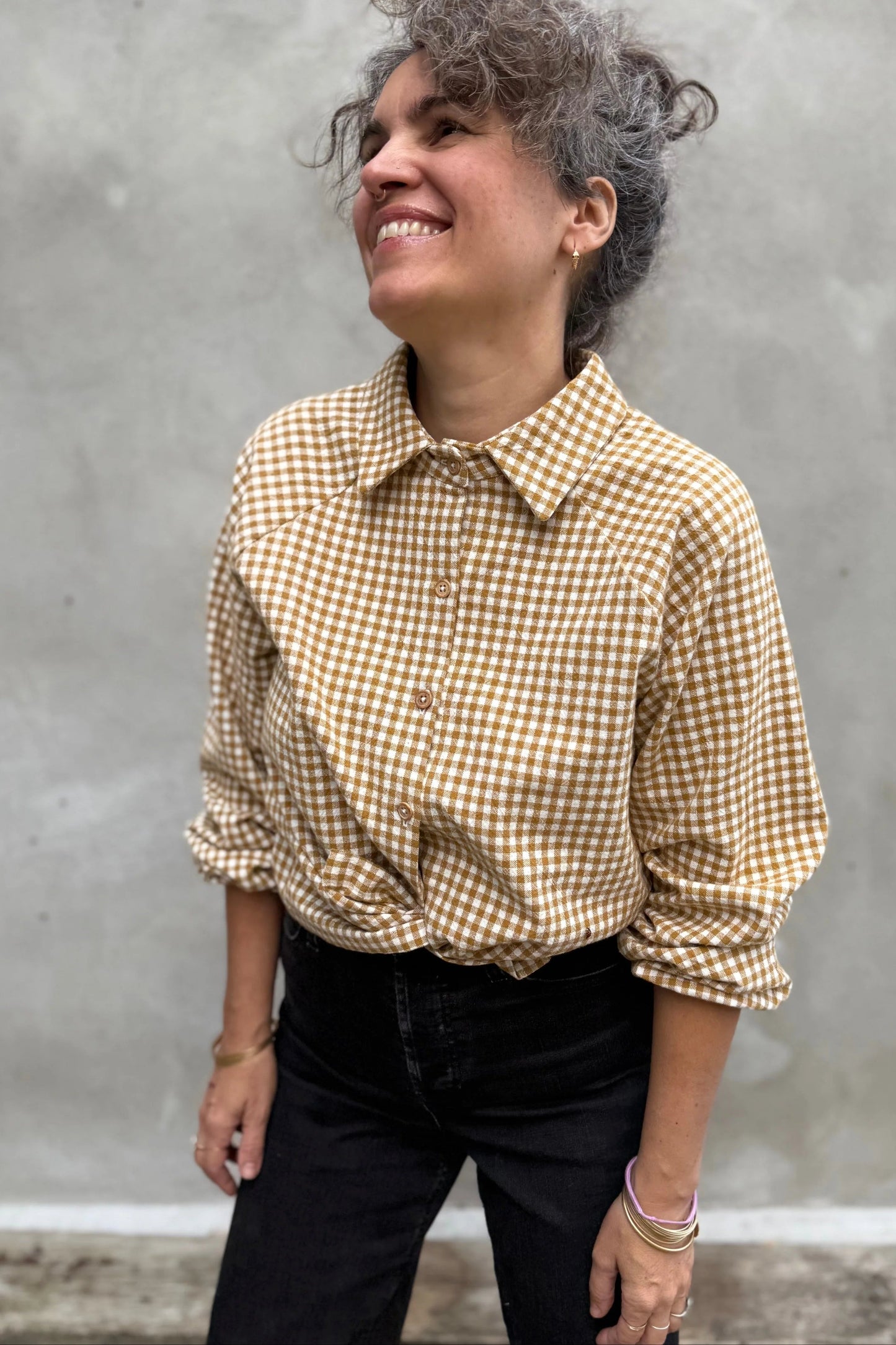 A woman wearing the Acer Blouse by Kazak in Ochre Gingham, a raglan sleeved blouse with a classic collar, button front, and elastic cuffs. She is wearing it with black jeans and standing in front of a grey wall.