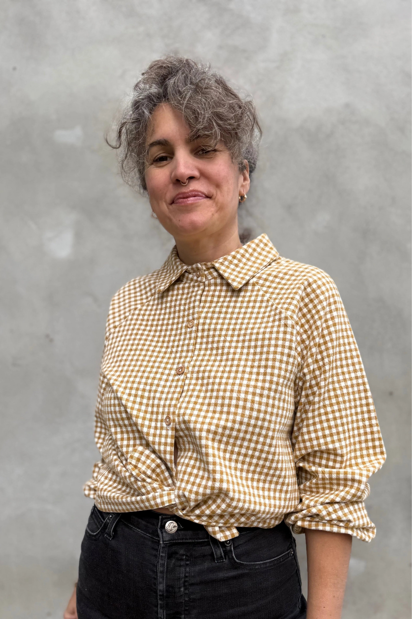A woman wearing the Acer Blouse by Kazak in Ochre Gingham, a raglan sleeved blouse with a classic collar, button front, and elastic cuffs. She is wearing it with black jeans and standing in front of a grey wall.