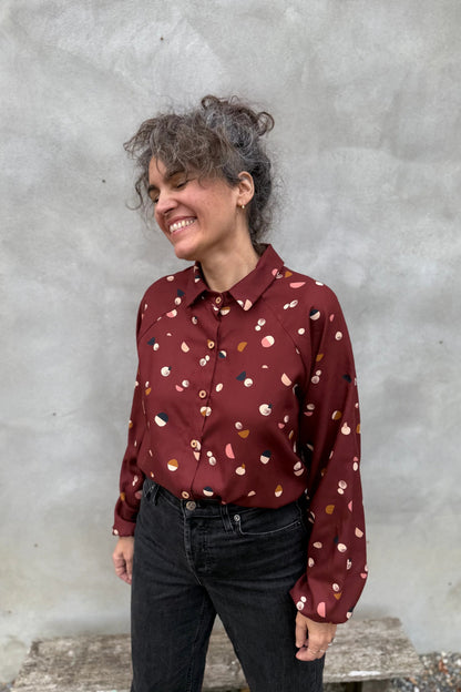 A woman wearing the Acer Blouse by Kazak in Burgundy Moons, a raglan sleeved blouse with a classic collar, button front, and elastic cuffs. She is wearing it with black jeans and standing in front of a grey background. 