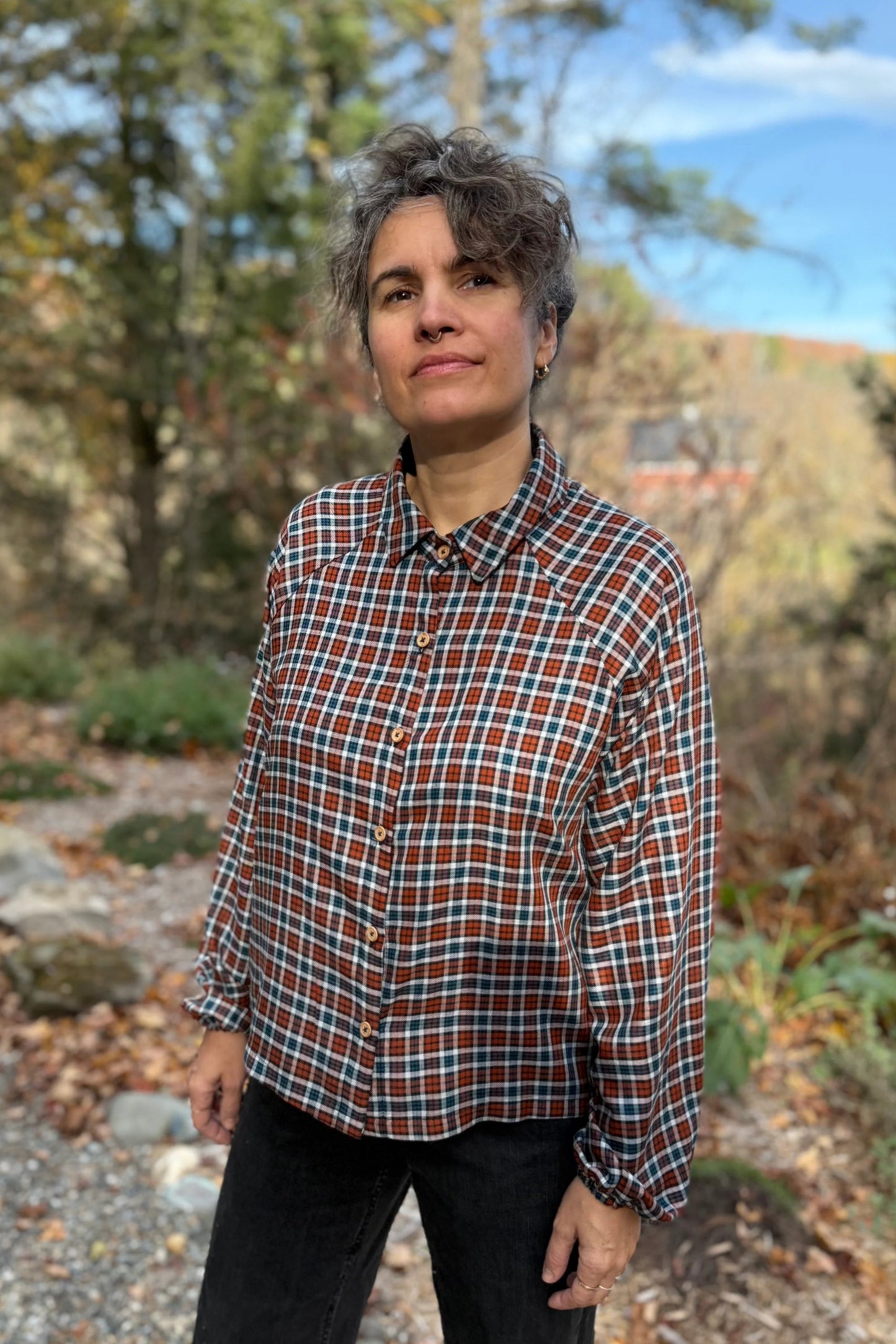 A woman wearing the Acer Blouse by Kazak in Checkered fabric, a raglan sleeved blouse with a classic collar, button front, and elastic cuffs. She is wearing it with black jeans and standing in a forest.