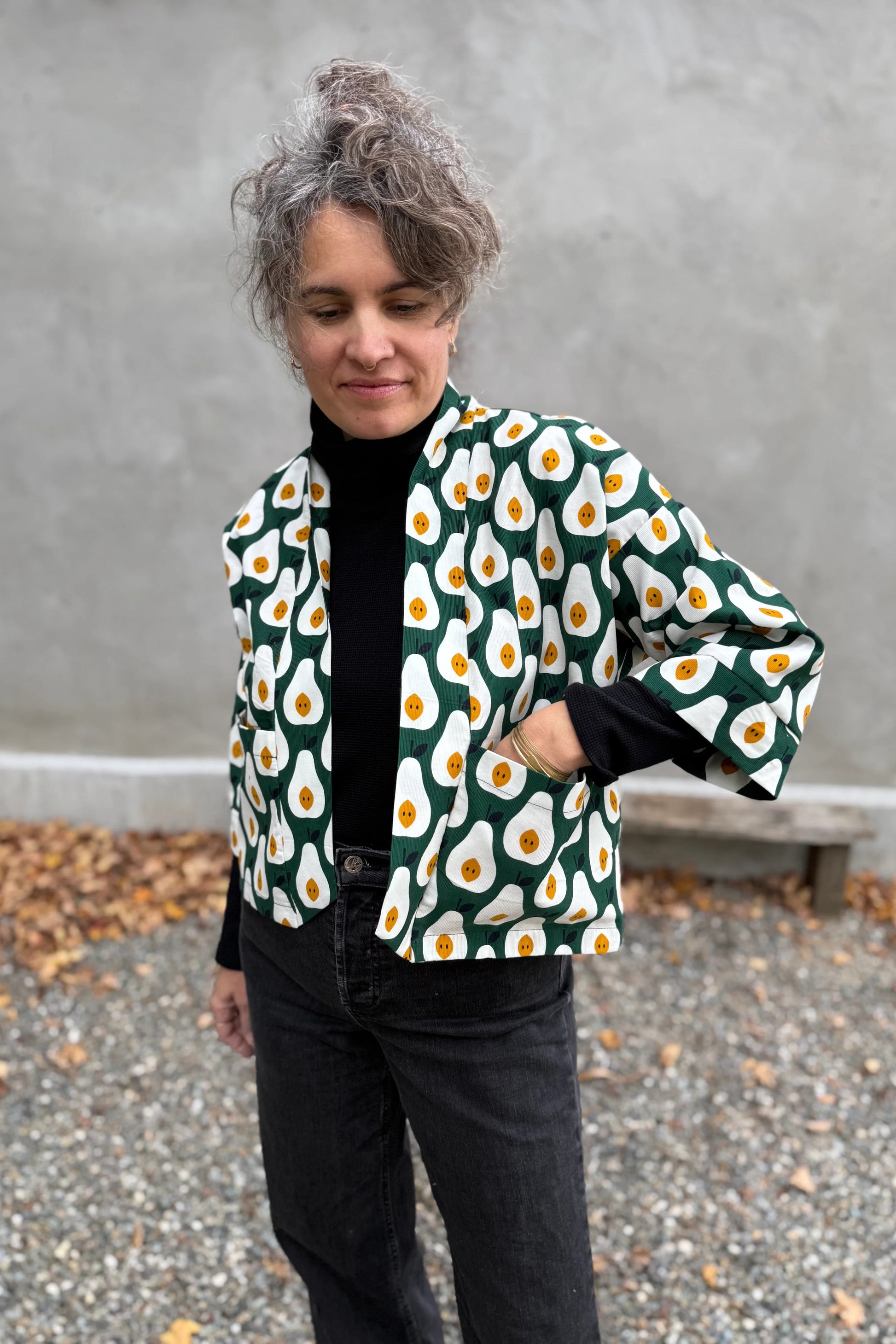 A woman wearing the Magnolia Jacket by Kazak in green and white corduroy Pears print fabric, a short kimono style open jacket with 3/4 sleeves and large patch pockets. She is wearing it with a black top and black jeans and standing in front of a grey wall. 