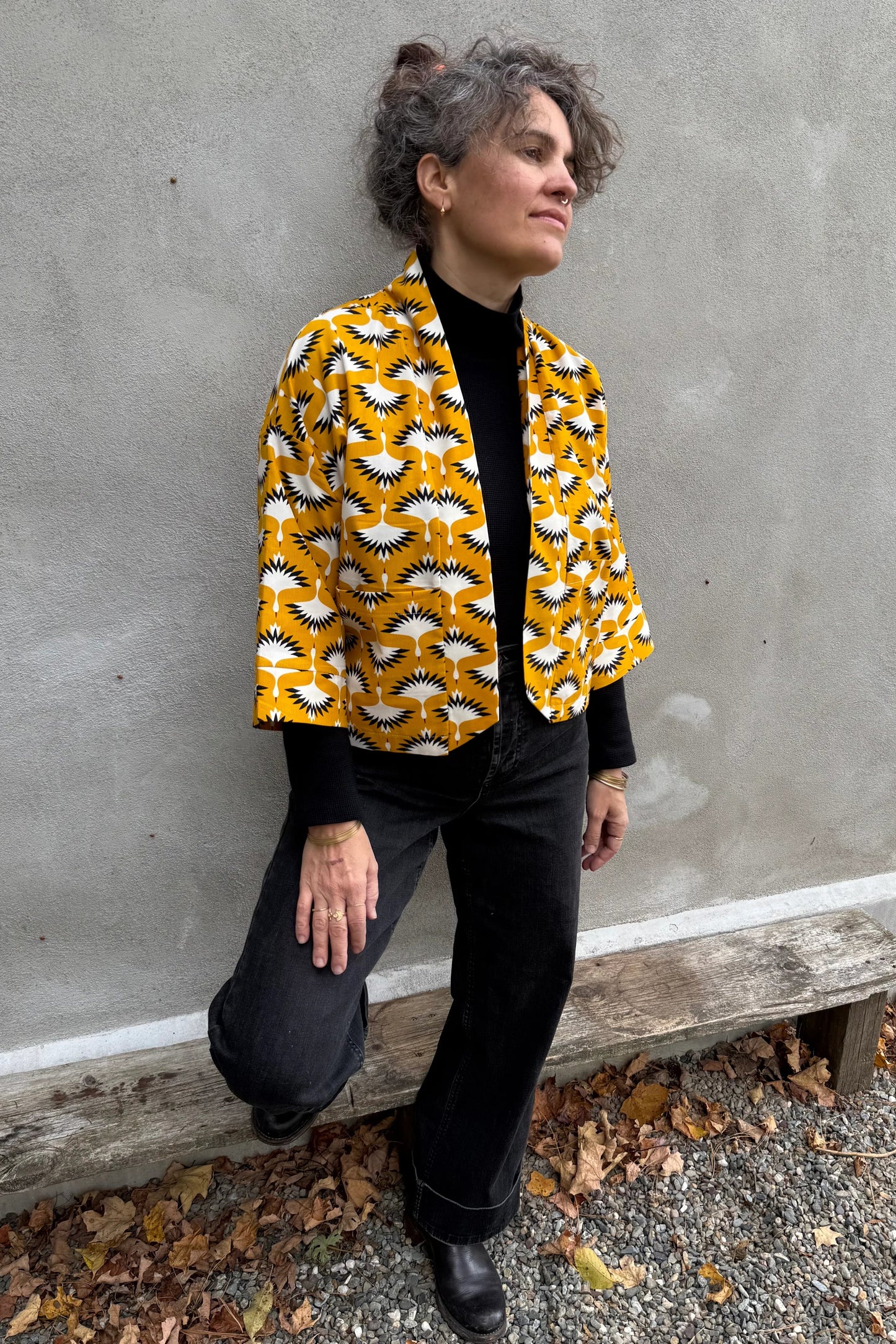 A woman wearing the Magnolia Jacket by Kazak in yellow corduroy Cranes print fabric, a short kimono style open jacket with 3/4 sleeves and large patch pockets. She is wearing it with a black top and black jeans and standing in front of a grey wall. 
