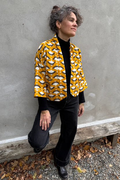 A woman wearing the Magnolia Jacket by Kazak in yellow corduroy Cranes print fabric, a short kimono style open jacket with 3/4 sleeves and large patch pockets. She is wearing it with a black top and black jeans and standing in front of a grey wall. 