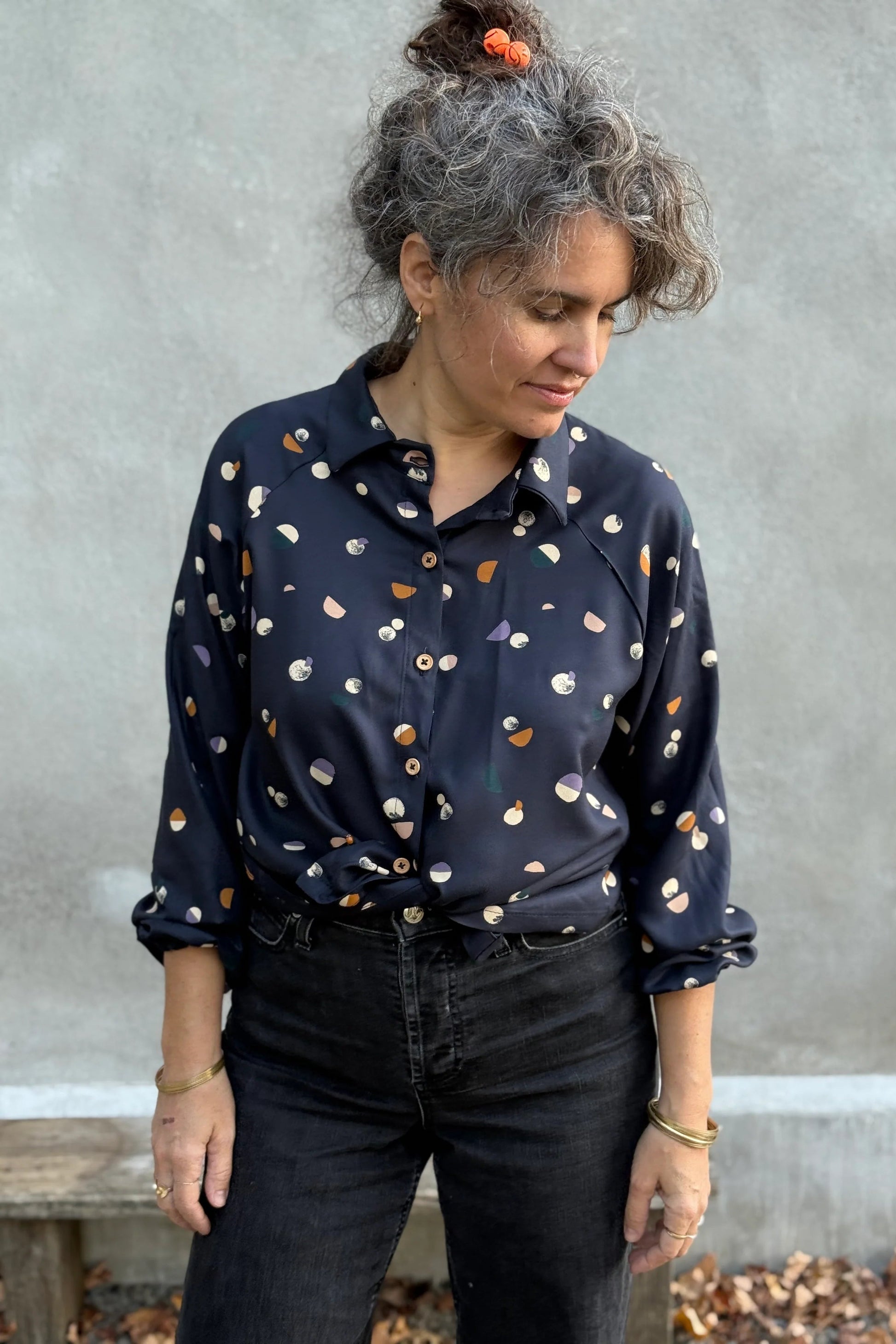 A woman wearing the Acer Blouse by Kazak in Midnight Moons, a raglan sleeved blouse with a classic collar, button front, and elastic cuffs. She is wearing it with black jeans and standing in front of a grey wall. 