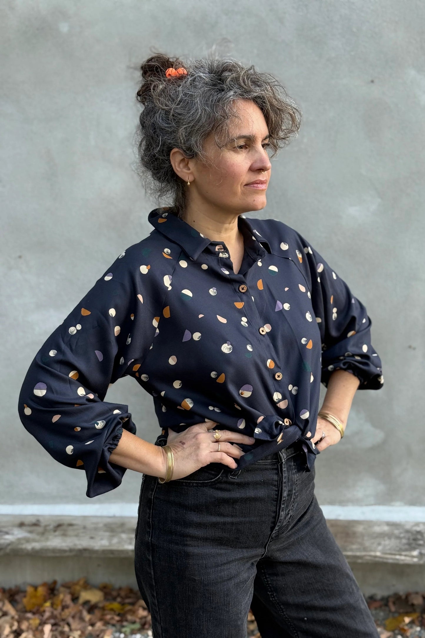 A woman wearing the Acer Blouse by Kazak in Midnight Moons, a raglan sleeved blouse with a classic collar, button front, and elastic cuffs. She is wearing it with black jeans and standing in front of a grey wall. 