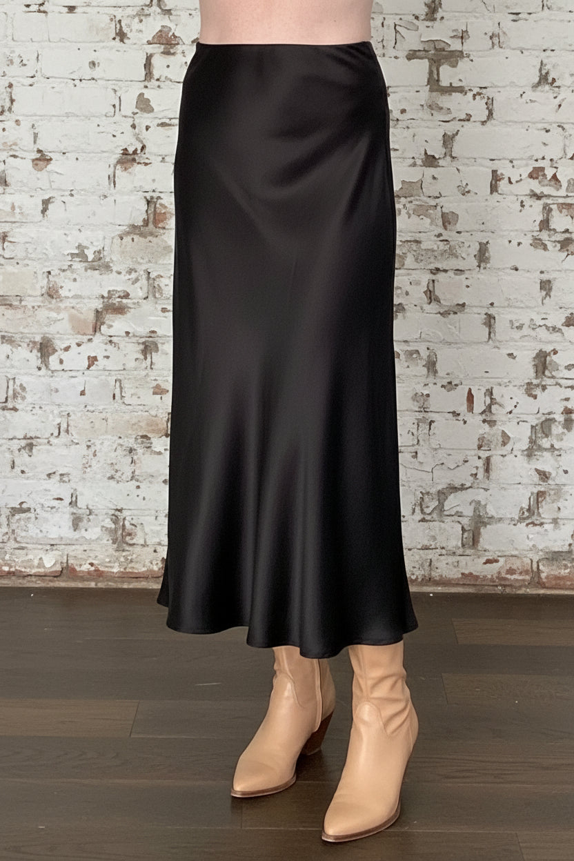 Waist down view of a woman wearing the Satin Skirt by Dotty in Black, a long bias cut A-line skirt with an elastic waistband. She is standing indoors in front of a white brick wall. 