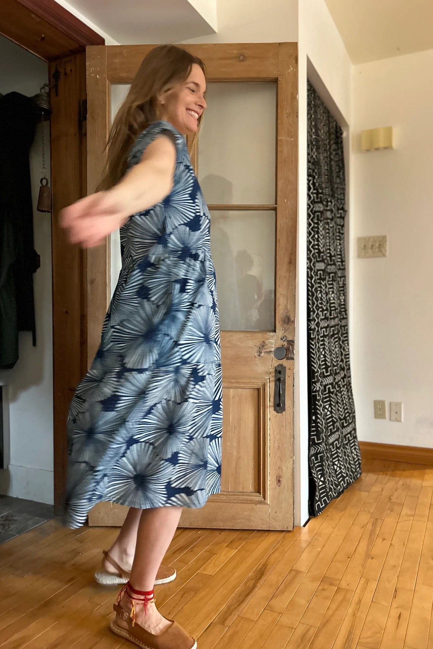 Side view of Woman wearing the Lavendula Dress by Kazak in Floral Indigo with cream, a short sleeved square neck midi dress with three ruffles. She is twirling in a room with wooden floors and a door.