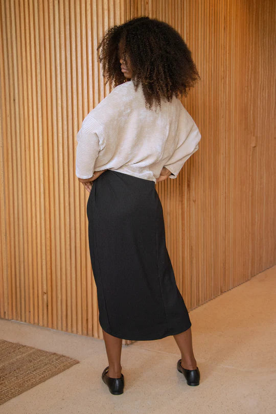 A woman wearing the Amsterdam Sweater by MAS in Latte, an oversized one-size top with a chimney collar, extended sleeves, and a cropped length, with blue trousers, standing in front of a wooden wall. 