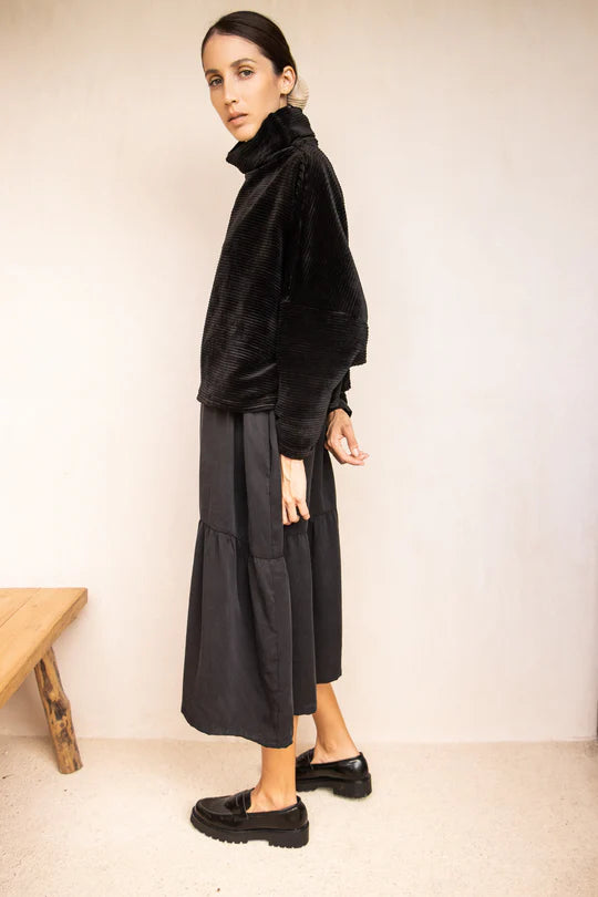 Side view of a woman wearing the Amsterdam Sweater by MAS in Pepper, an oversized one-size top with a chimney collar, extended sleeves, and a cropped length, with a black skirt,  standing in front of a beige wall. 