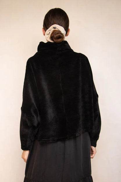 Back view of a woman wearing the Amsterdam Sweater by MAS in Pepper, an oversized one-size top with a chimney collar, extended sleeves, and a cropped length, with a black skirt,  standing in front of a beige wall. 