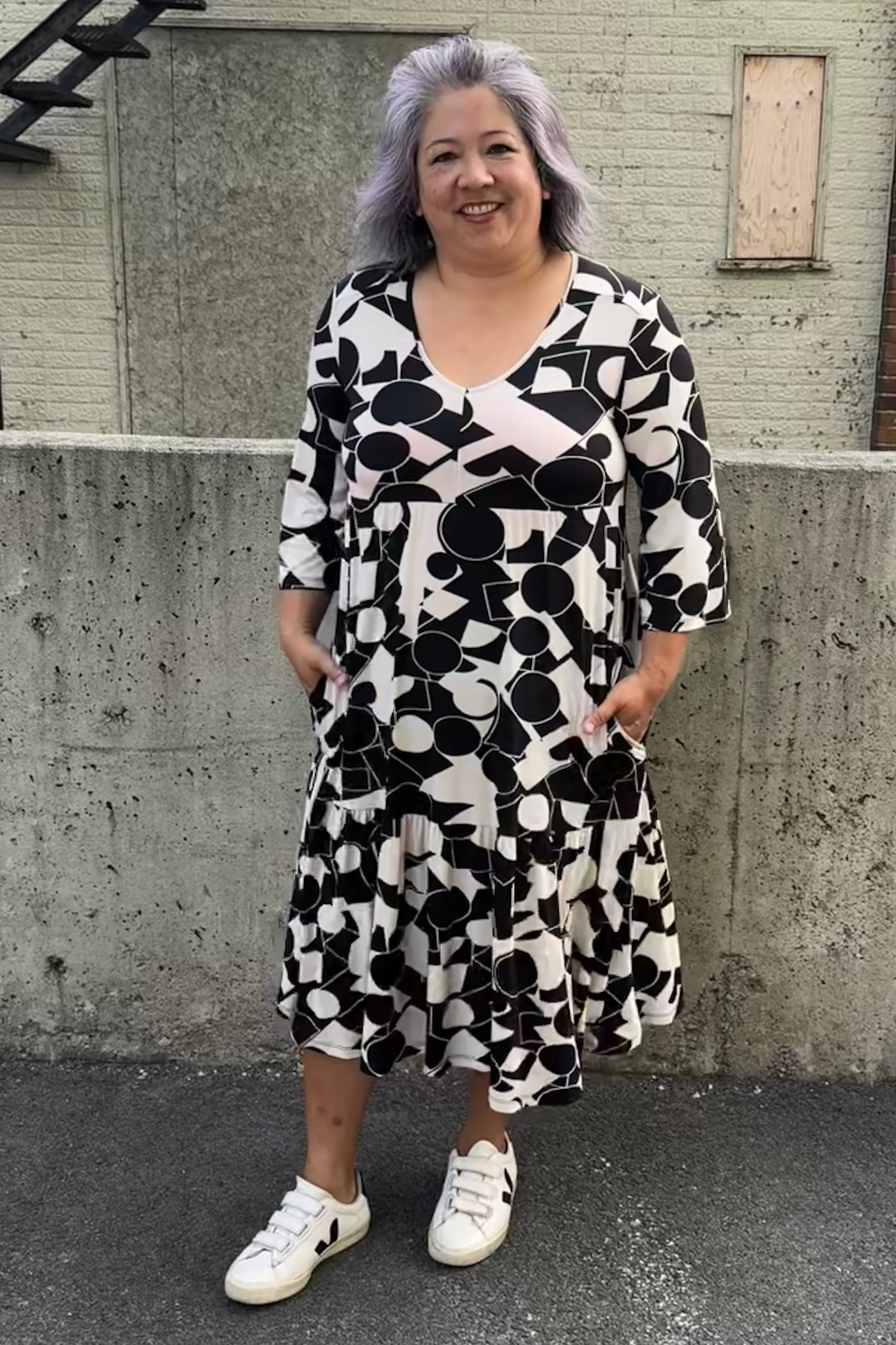 A woman wearing the Farah Dress by Pure Essence in Black/Ivory, a geometric print dress with a v-neck, 3/4-sleeves, baby doll shape, tiered midi length skirt, and front patch pockets. She is standing in front of a brick building. 