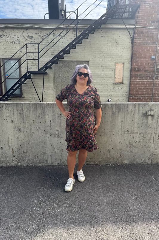 A woman wearing the Botanical Dress by Karkass in Floral, a knit dress with a V-neck, elbow length sleeves, fit and flare shape, and above the knee hem.  She is standing in front of a building with a fire escape.  