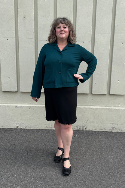 A woman wearing the Fern Jacket by Pure Essence in Spruce, a boxy jacket with a lapel collar, button front and patch pockets in a ribbed fabric. She is wearing it with a black skirt and standing in front of a grey wall. 