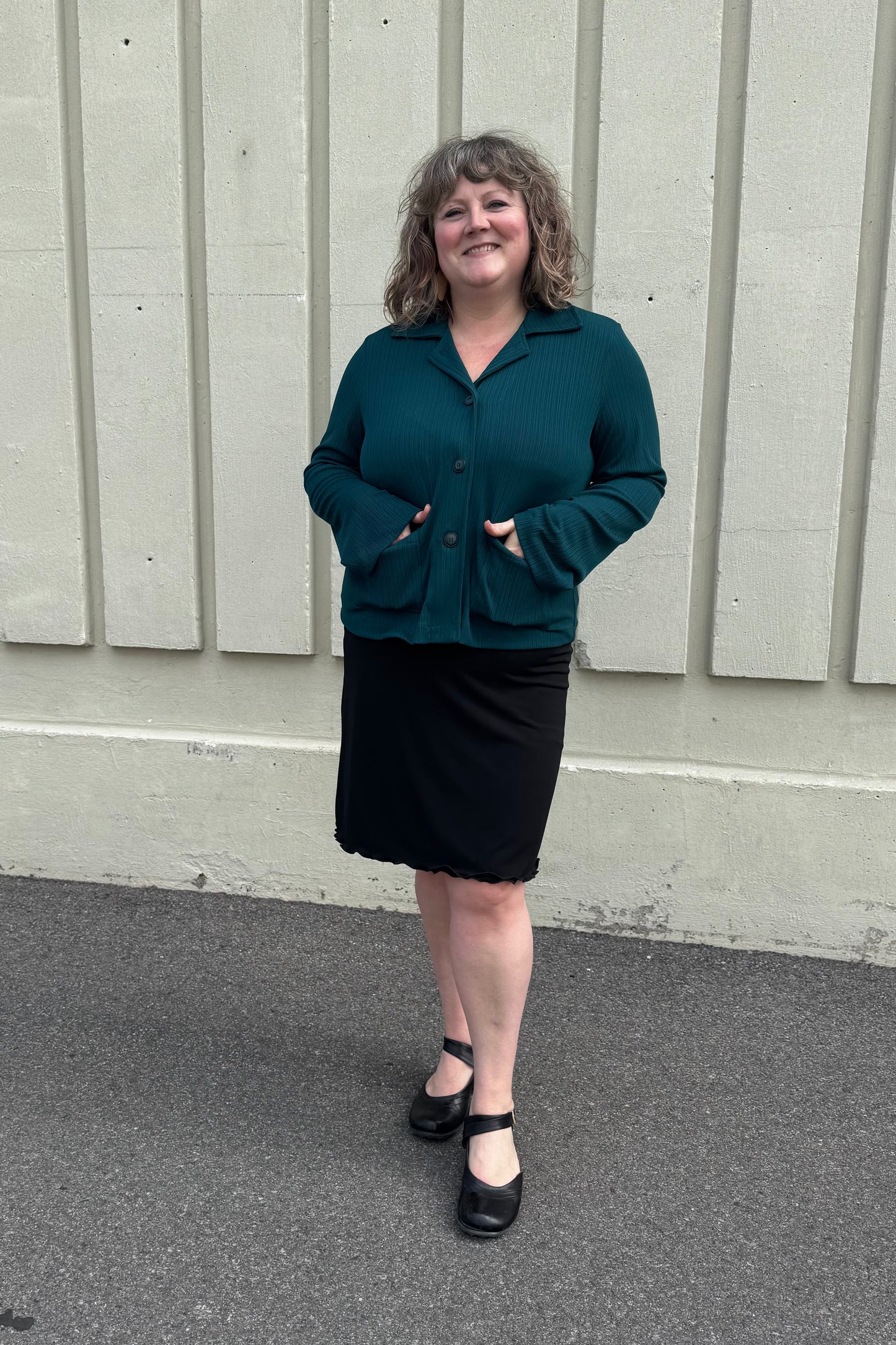 A woman wearing the Fern Jacket by Pure Essence in Spruce, a boxy jacket with a lapel collar, button front and patch pockets in a ribbed fabric. She is wearing it with a black skirt and standing in front of a grey wall. 