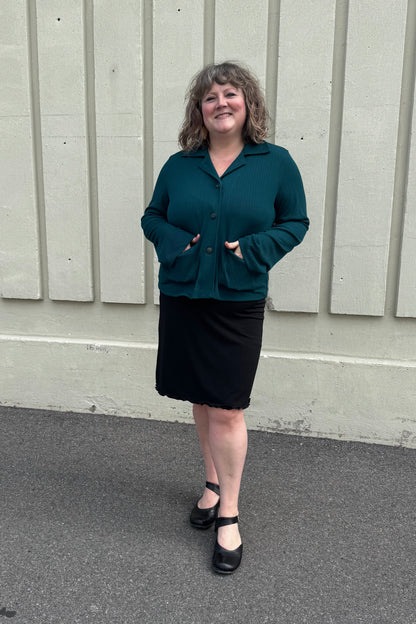 A woman wearing the Fern Jacket by Pure Essence in Spruce, a boxy jacket with a lapel collar, button front and patch pockets in a ribbed fabric. She is wearing it with a black skirt and standing in front of a grey wall. 