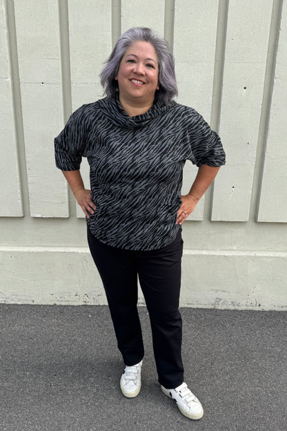 A woman wearing the Francis Tunic by Pure in Black/Charcoal animal print, featuring a turtleneck, 3/4 sleeves and two patch pockets. She is wearing it with Black pants and standing in front of a grey wall. 
