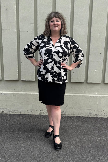 A woman wearing the Francesca Blouse by Pure in Black/Ivory geometric print, an oversized blouse with a lapel collar, button front, 3/4 sleeves with button and tab detail, and rounded h-low hemline. She is wearing it with a black skirt and standing against a grey wall.