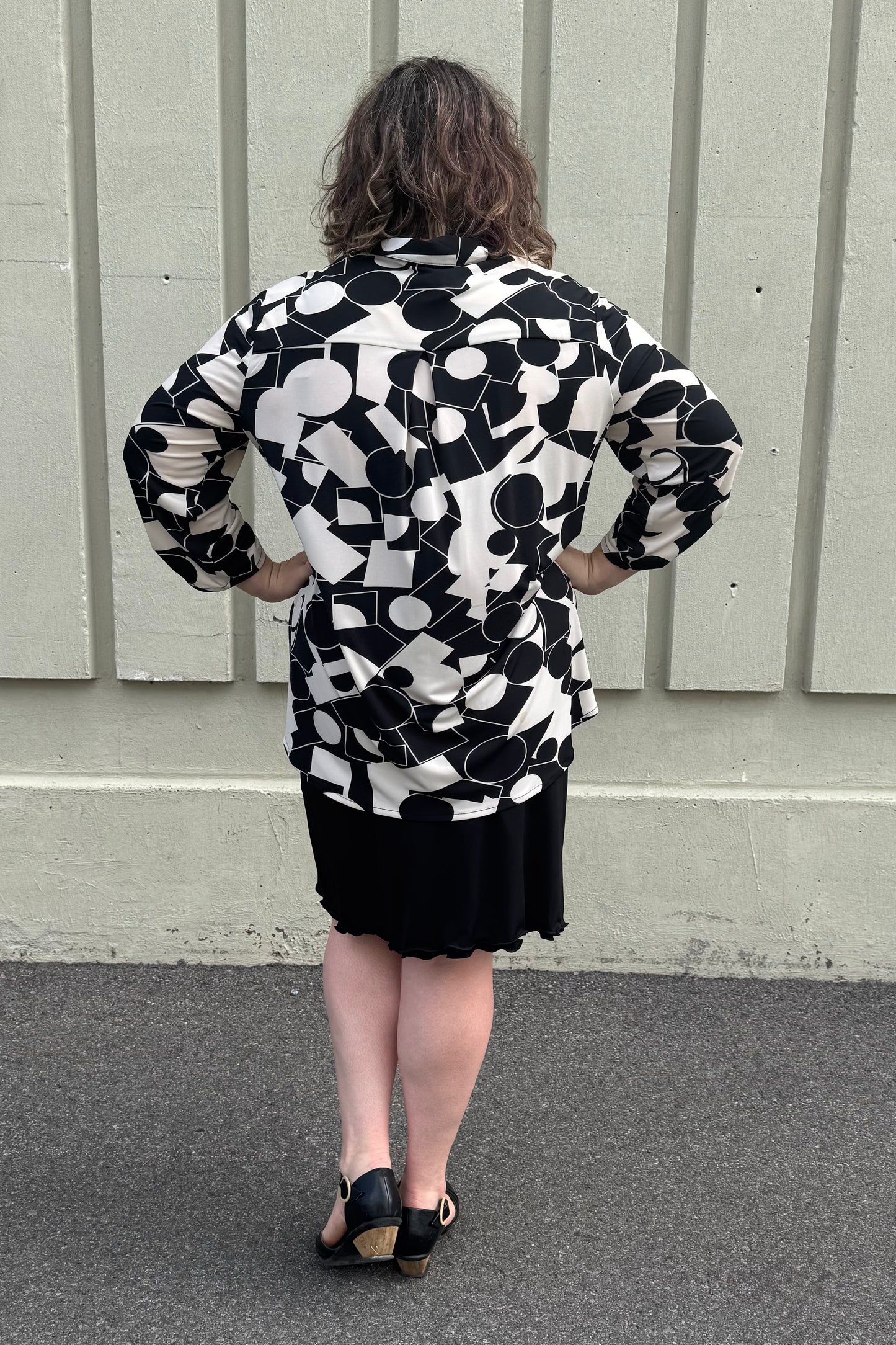 Back view of a woman wearing the Francesca Blouse by Pure in Black/Ivory geometric print, an oversized blouse with a lapel collar, button front, 3/4 sleeves with button and tab detail, and rounded h-low hemline. She is wearing it with a black skirt and standing against a grey wall.