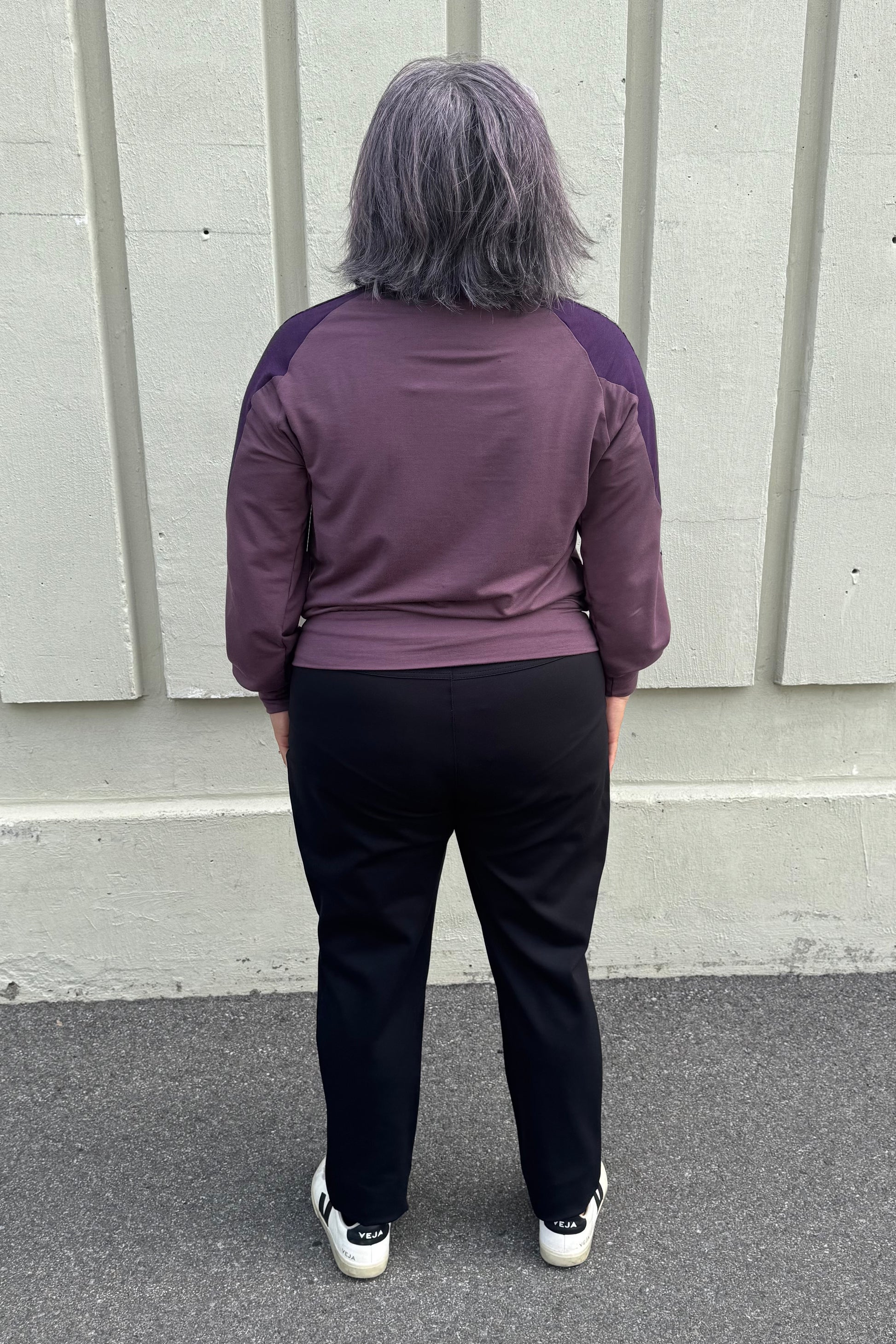 Back view of a woman wearing the Erin Sweater by Solomia in Cream/Black/Purple, a colour blocked top with a round neck and long sleeves, and a mix of patterns an solid colours including purple leopard print. She is wearing it with black pants and standing outdoors in front of a grey wall. 