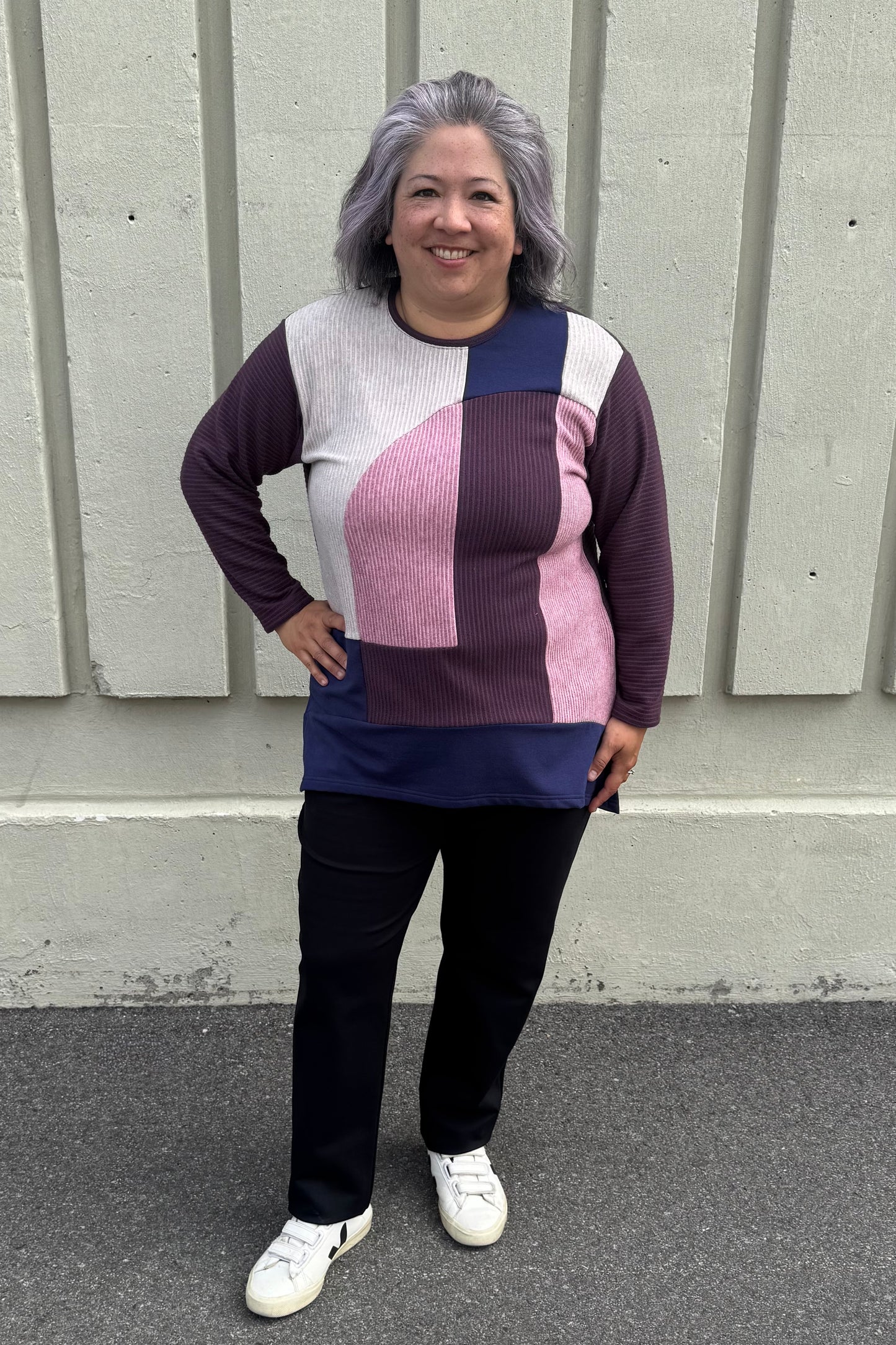 A woman wearing the Gladis Sweater by Solomia in Burgundy Pink Navy, a colour blocked in a mix of textures with a round neck, long sleeves, below the hip length ad side slits. She is wearing it with black pants and standing outside in front of a grey wall. 