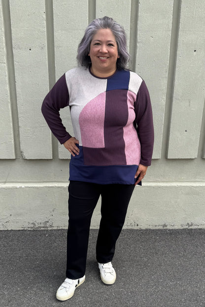A woman wearing the Gladis Sweater by Solomia in Burgundy Pink Navy, a colour blocked in a mix of textures with a round neck, long sleeves, below the hip length ad side slits. She is wearing it with black pants and standing outside in front of a grey wall. 
