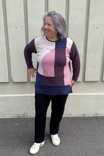A woman wearing the Gladis Sweater by Solomia in Burgundy Pink Navy, a colour blocked in a mix of textures with a round neck, long sleeves, below the hip length ad side slits. She is wearing it with black pants and standing outside in front of a grey wall. 