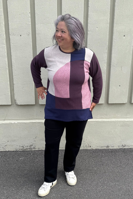 A woman wearing the Gladis Sweater by Solomia in Burgundy Pink Navy, a colour blocked in a mix of textures with a round neck, long sleeves, below the hip length ad side slits. She is wearing it with black pants and standing outside in front of a grey wall. 