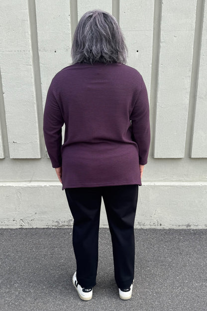 Back view of a woman wearing the Gladis Sweater by Solomia in Burgundy Pink Navy, a colour blocked in a mix of textures with a round neck, long sleeves, below the hip length ad side slits. She is wearing it with black pants and standing outside in front of a grey wall. 