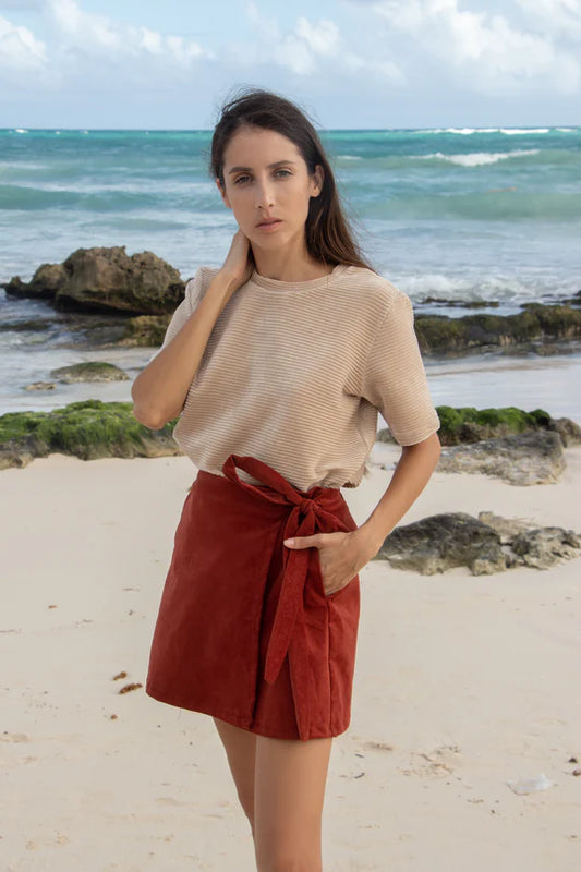 A woman wearing the MAS Tofino Top in Latte, a velvet rib t-shirt with a round neck, short sleeves, and a slightly cropped length. She is wearing it with a red skirt and standing on a beach in front of the ocean. 