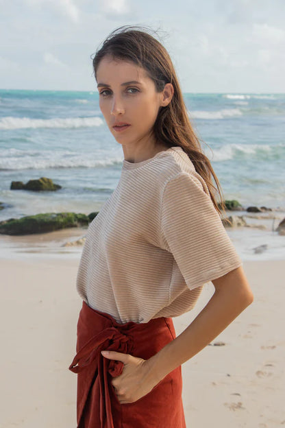 Side view of a woman wearing the MAS Tofino Top in Latte, a velvet rib t-shirt with a round neck, short sleeves, and a slightly cropped length. She is wearing it with a red skirt and standing on a beach in front of the ocean. 