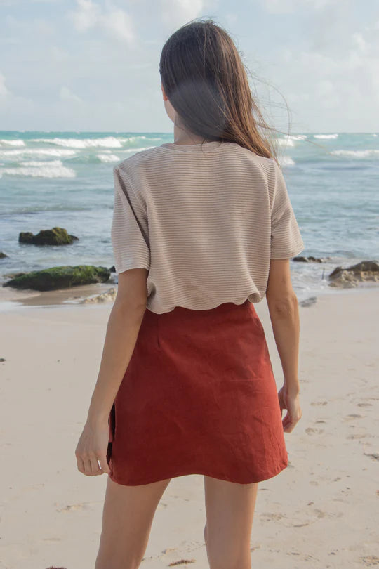 Back view of a woman wearing the MAS Tofino Top in Latte, a velvet rib t-shirt with a round neck, short sleeves, and a slightly cropped length. She is wearing it with a red skirt and standing on a beach in front of the ocean. 