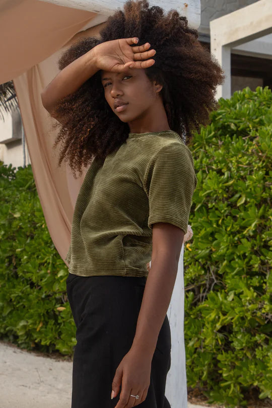 Side view of a woman wearing the MAS Tofino Top in Moss, a velvet rib t-shirt with a round neck, short sleeves, and a slightly cropped length. She is wearing it with black pants and standing in front of a bush. 