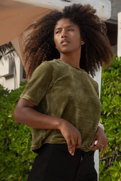 A woman wearing the MAS Tofino Top in Moss, a velvet rib t-shirt with a round neck, short sleeves, and a slightly cropped length. She is wearing it with black pants and standing in front of a bush. 