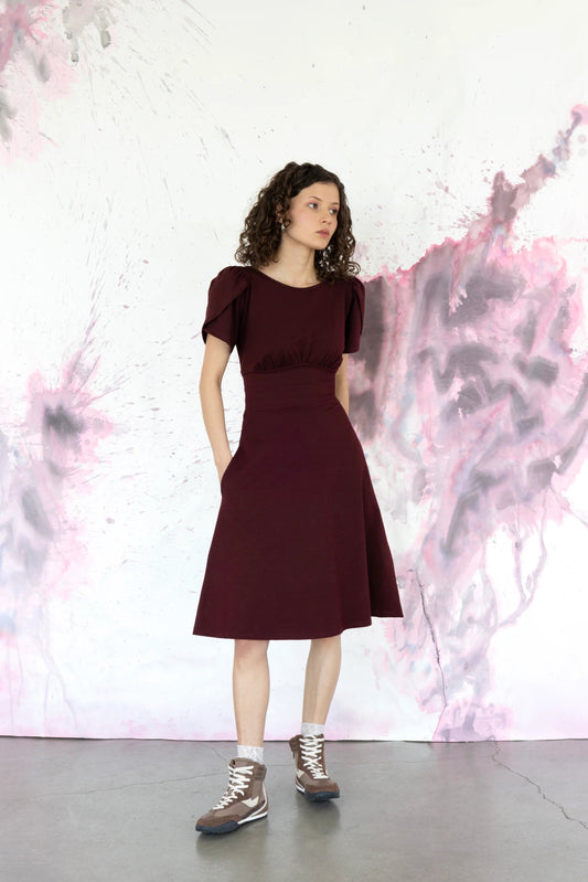 A woman wearing the Tulipe dress by Eve Lavoie in Burgundy, with short puffed tulip sleeves, a pleated bodice with a wide waistband and attached tie belt, and knee-length A-line skirt. It has a round neck on one side and scoop neck on the other and is reversible back to front. She is standing against a colourful abstract background. 