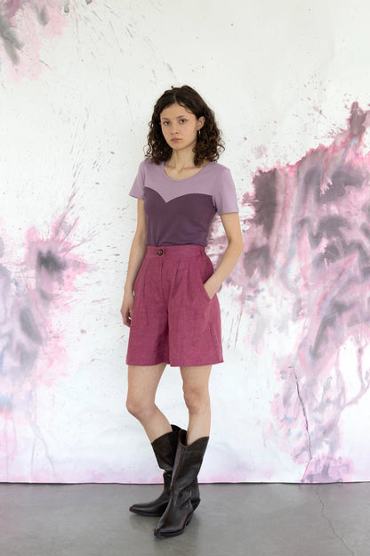 A woman wearing the Aster Shorts by Eve Lavoie in Raspberry Stripe with an 8-inch inseam, double pleats, a zip fly, an elasticated waistband and side pockets. She is wearing them with the Gentiane Top in Grape/Lilac and standing in front of a purple and white speckled background.
