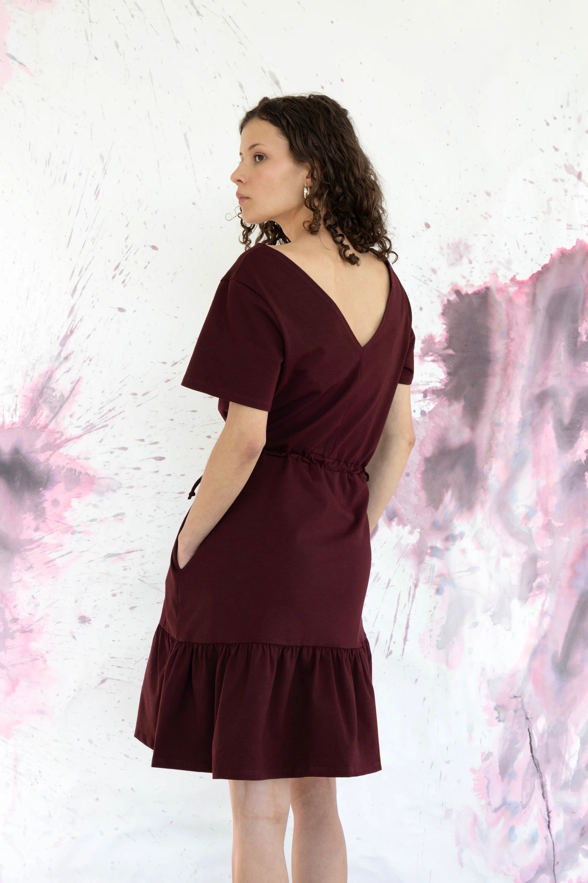 Back view of a woman wearing the Épervière Dress by Eve Lavoie in Fuchsia, a reversible dress with short sleeves, a V-neck on one side and a round neck on the other, a flounced hem, and ties at the sides of the waists. She is standing in front of a purple and grey abstract wall. 