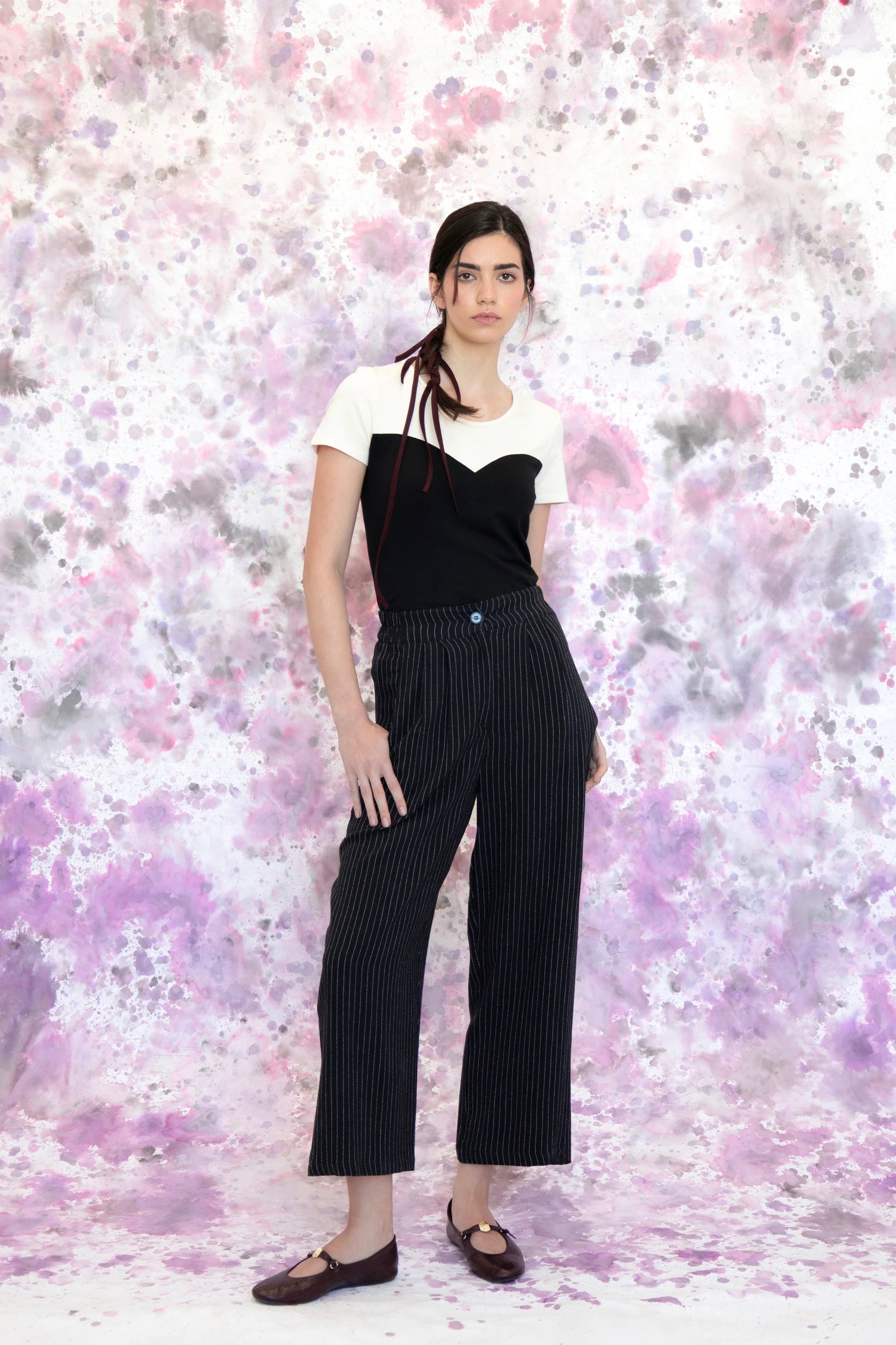 A woman wearing the Tanaisie Woven Pants from Eve Lavoie in Black Stripe, slightly cropped linen/cotton pants with wide legs, an elastic waist, classic fly closure, and slanted side pockets. She is wearing them with a black and white top and standing in front of a purple and white speckle background. 
