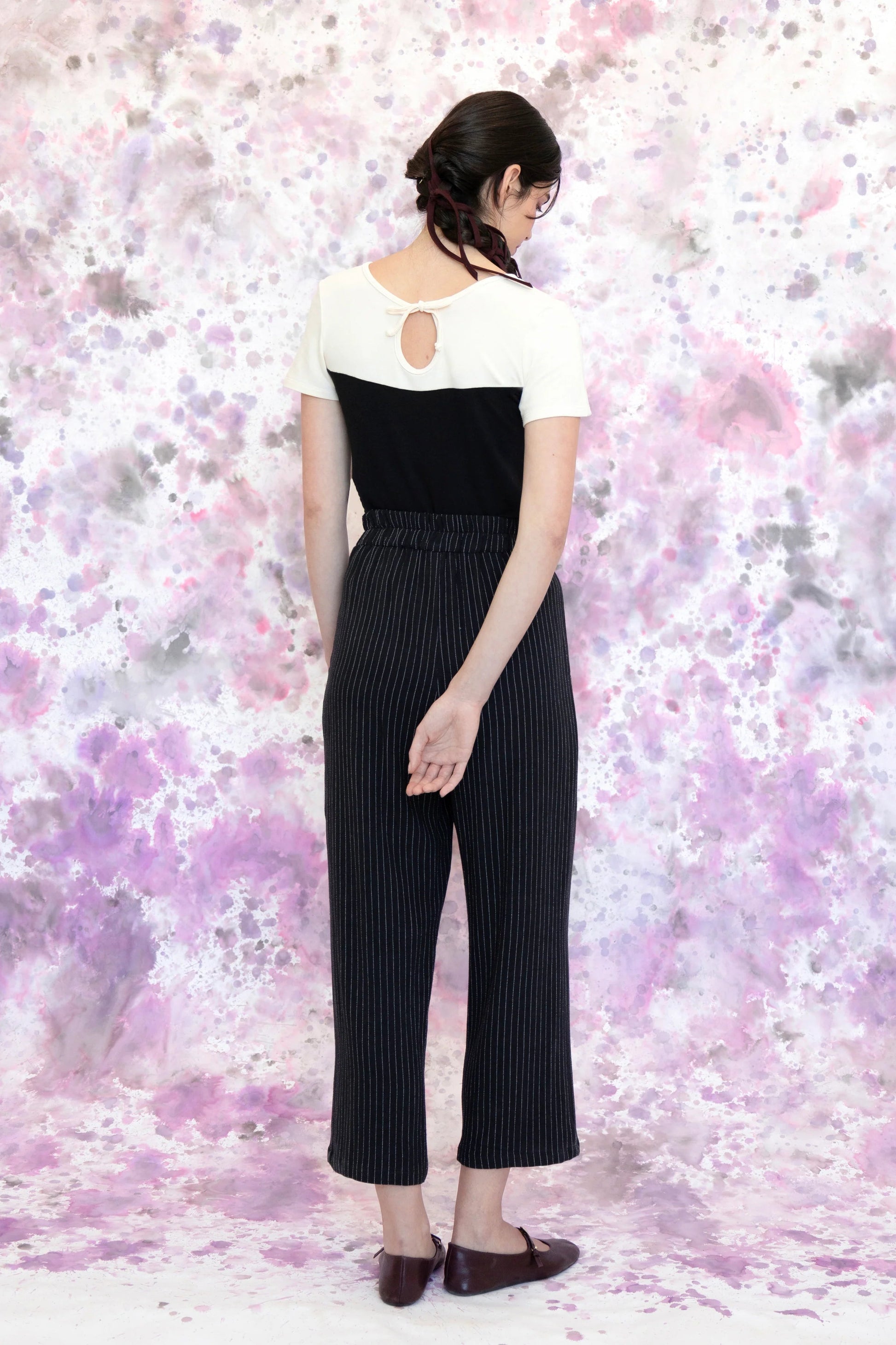 Back view of a woman wearing the Tanaisie Woven Pants from Eve Lavoie in Black Stripe, slightly cropped linen/cotton pants with wide legs, an elastic waist, classic fly closure, and slanted side pockets. She is wearing them with a black and white top and standing in front of a purple and white speckle background. 