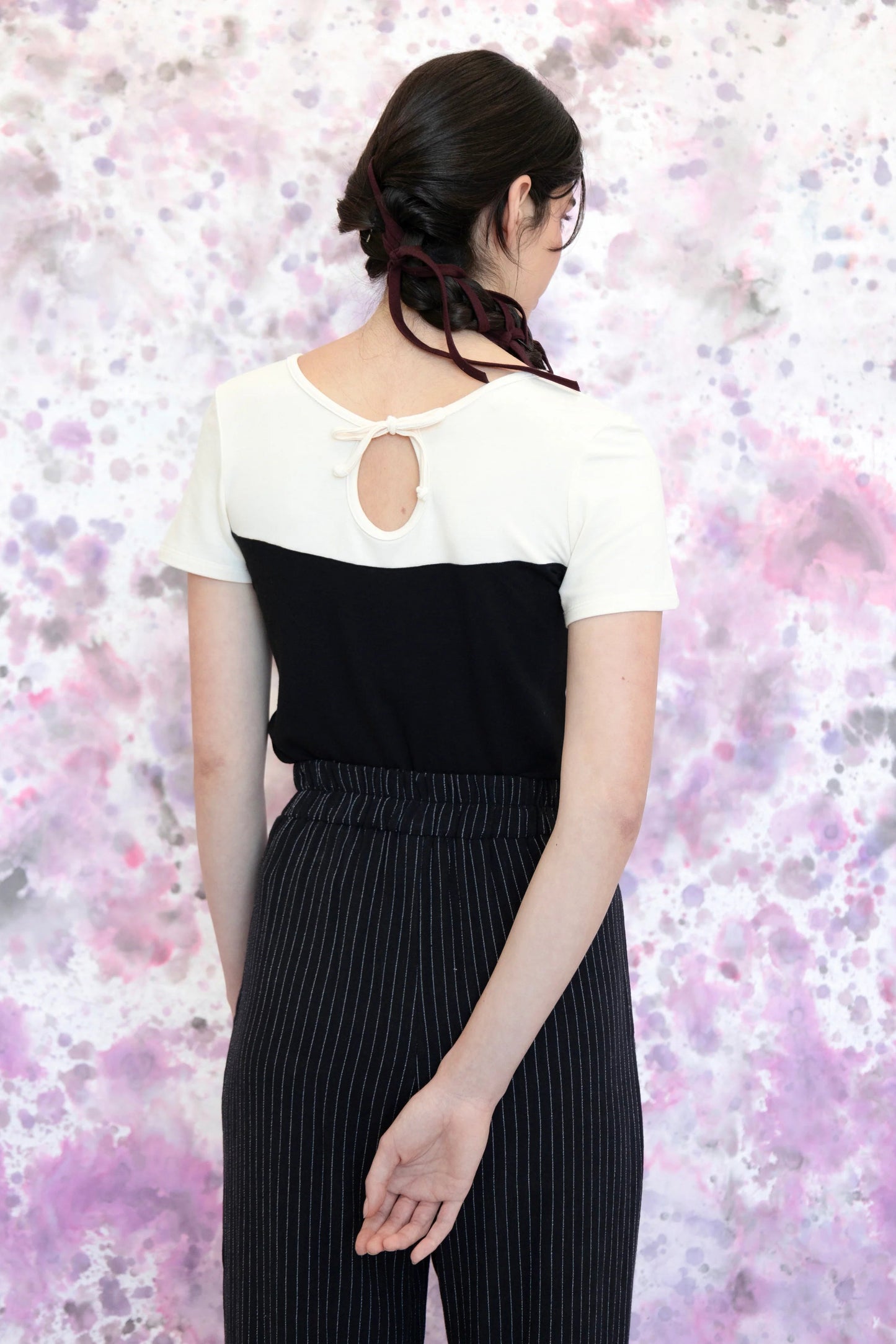 Back view of a woman wearing the Gentiane Top by Eve Lavoie in Black/White, a two-toned fitted tee with a sweetheart-effect neckline on one side and a cutout and tie detail on the other. She is wearing it with pinstripe pants and standing in front of a purple and grey abstract background. 