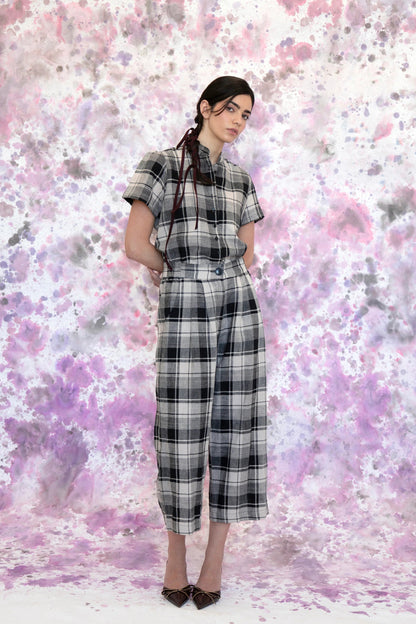 A woman wearing the Tanaisie Woven Pants from Eve Lavoie in Black and Cream Plaid, slightly cropped linen/cotton pants with wide legs, an elastic waist, classic fly closure, and slanted side pockets. She is wearing them with a black and white top and standing in front of a purple and white speckle background. 