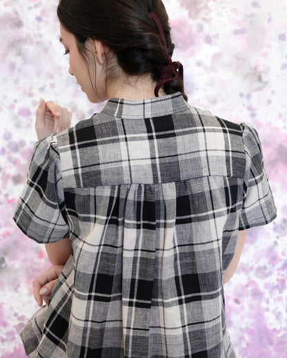 Back waist up view of a woman wearing the Eglantine Plaid Blouse by Eve Lavoie in Black and Cream Plaid, with a mandarin collar, short puffed sleeves, rounded hem, and button front. She is wearing it with matching pants and standing in front of a purple and white speckled background. 