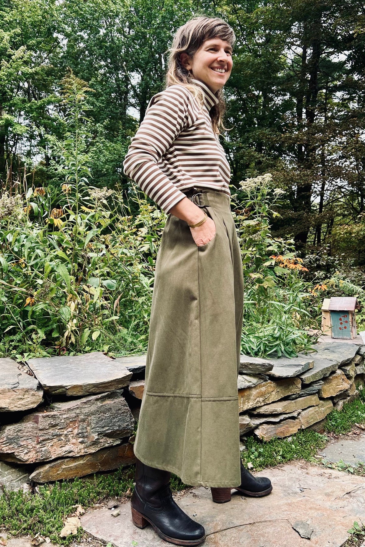 A woman standing in a lush garden wearing a black and white striped short-sleeve top, Kazak's Asarum skirt in Olive – a high-waisted midi skirt – and brown leather boots.