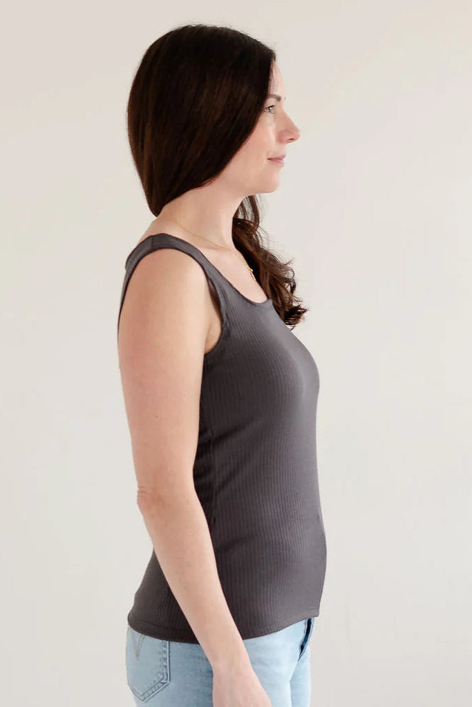 Side view of a woman wearing the Ang Hill Tracey Tank in Charcoal with wide straps, ribbed fabric, and a square neckline. She is wearing it with jeans and standing in a white room. 