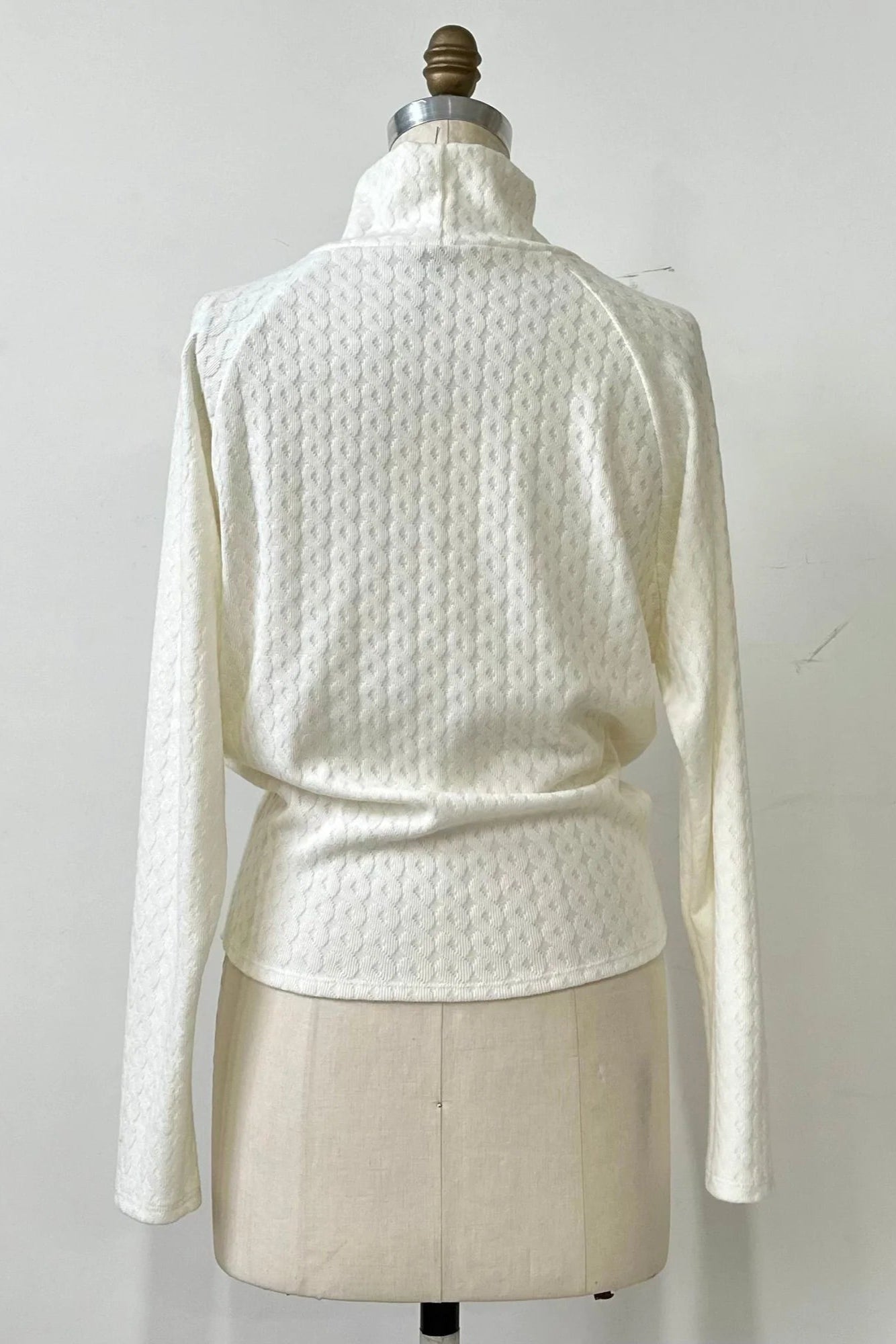 Back view of the Plume Sweater by Kollontai in White, a cable knit turtleneck with long tapered sleeves and a narrow hem for a bloused look, is shown on a mannequin in front of a white background. 