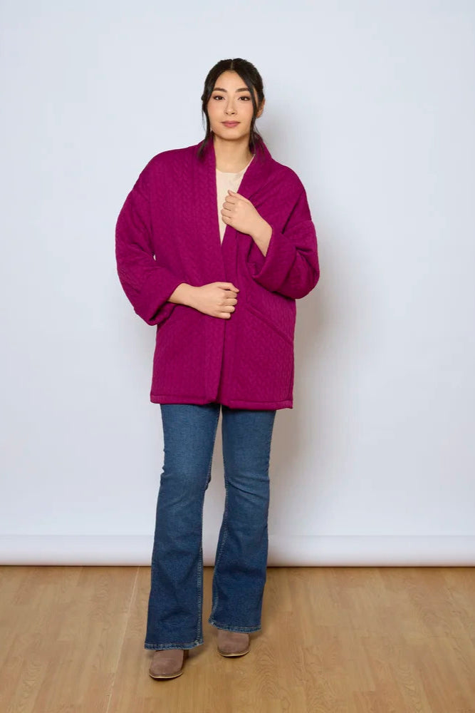 A woman wearing the Ina Coatigan by Tangente in Magenta, an open front cardigan in a cable-knit textured fabric, with dropped sleeves and patch pockets. She is wearing it with a white shirt and jeans and standing in front of a white background. 